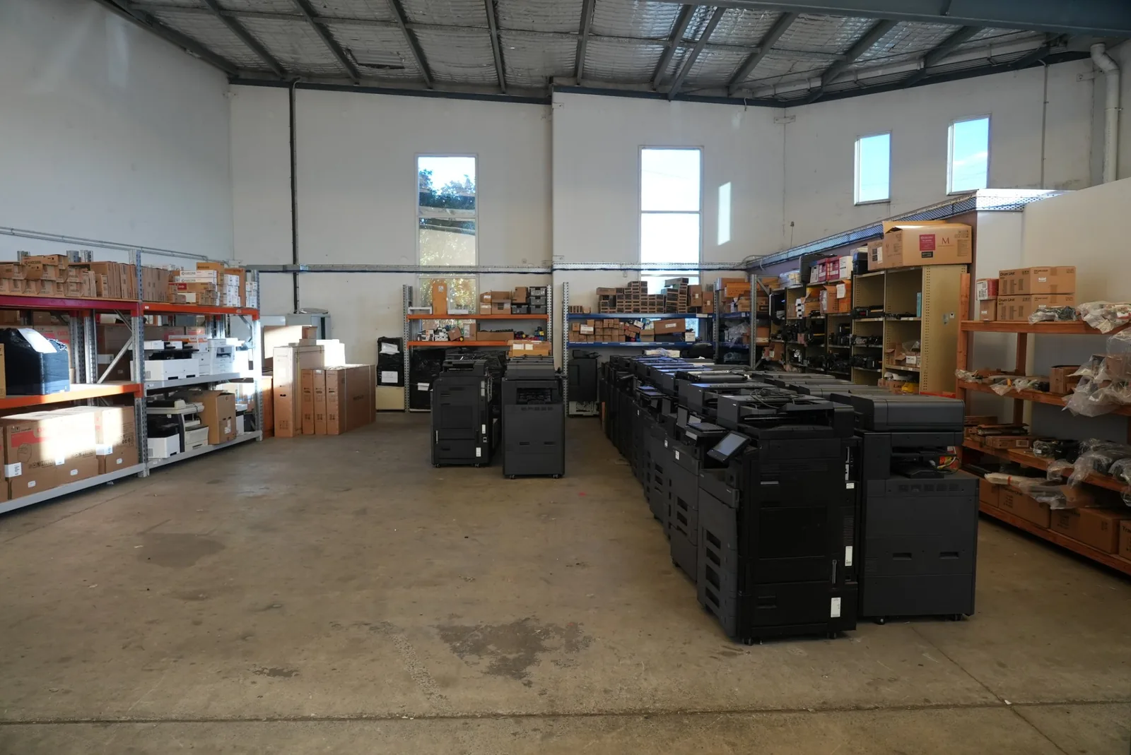 Wide view of the warehouse floor showing the printer fleet on the right and racked consumables and spare parts along both walls