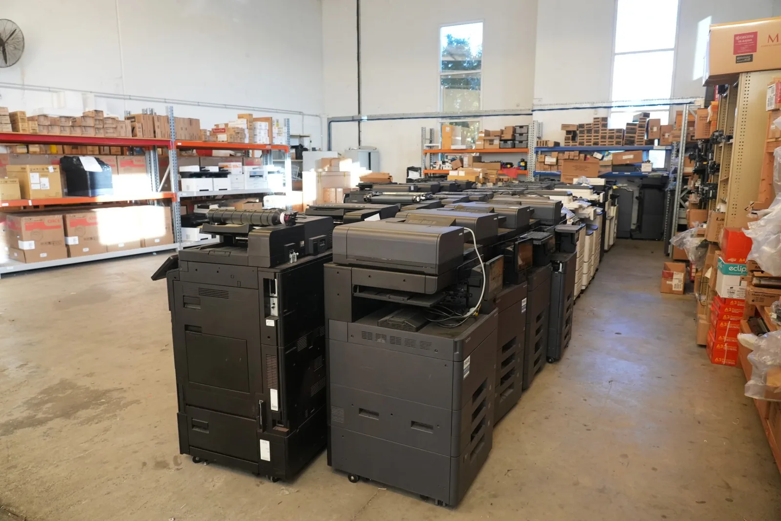 Rows of black multifunction office printers on the warehouse floor, with shelving of boxed toner and spare parts behind them