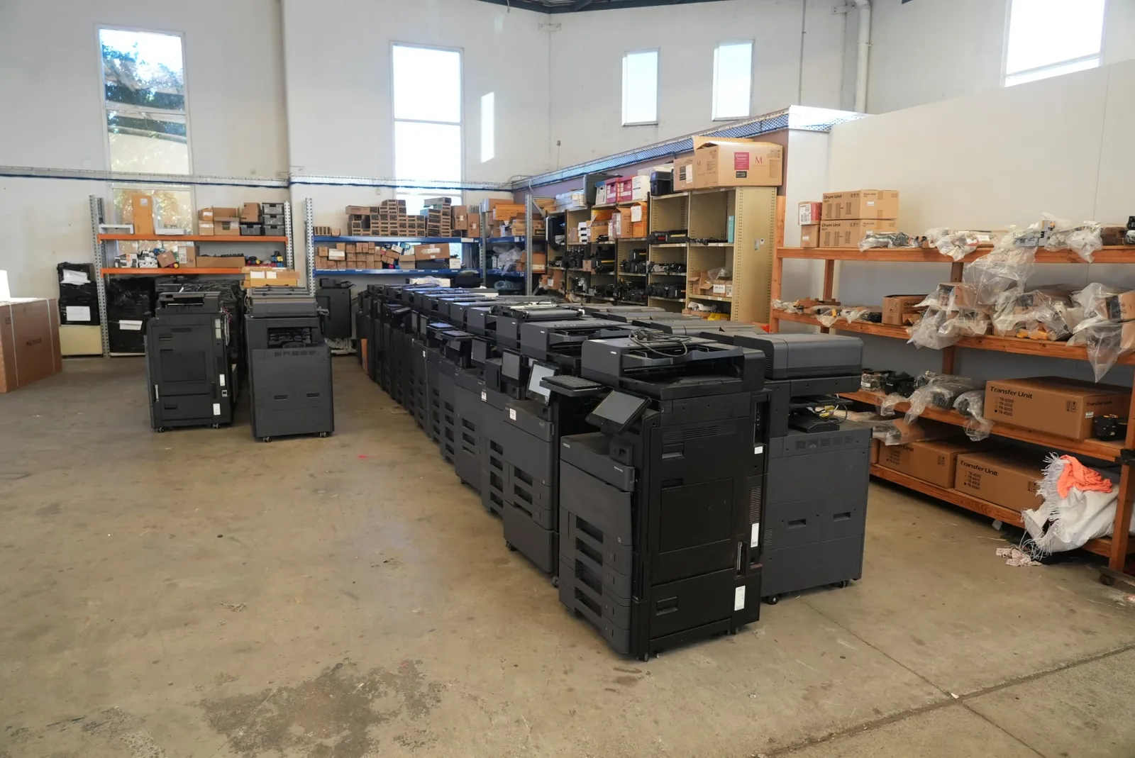 A long row of multifunction printers beside wall racking holding boxed drum units, transfer units and bagged service parts